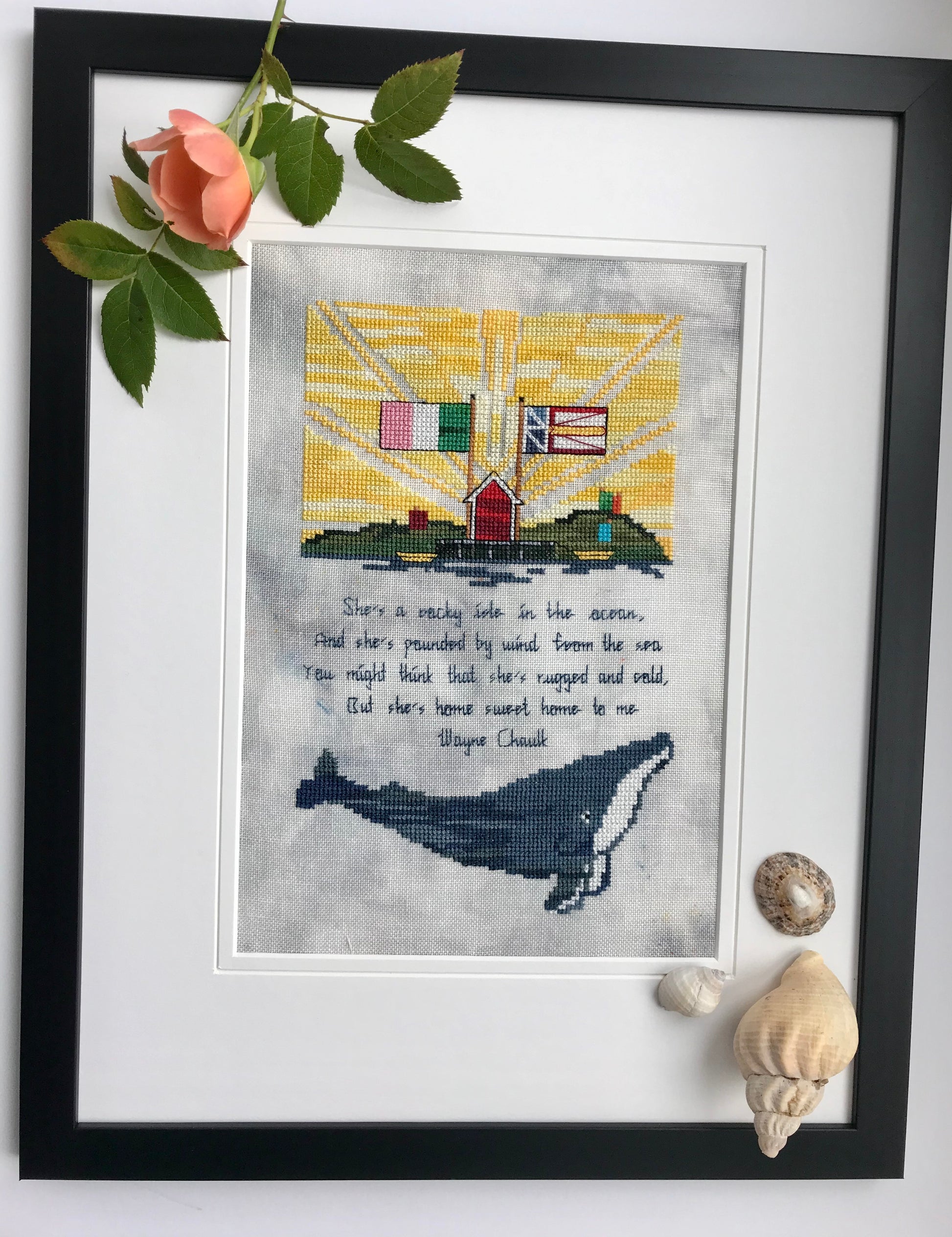Black wooden frame with a white mat and a real peach rose at the left corner on top of a cross stitched piece. Three different types of shells are on top of the mat on the bottom right. Cross stitched picture in the middle depicts two flags, a fishing stage, green hills, a song verse and a humpback whale.