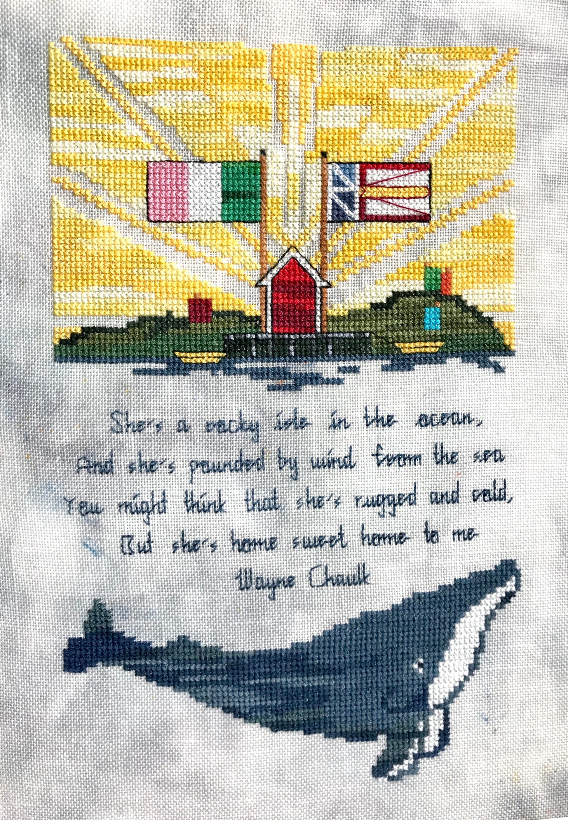 Cross stitched yellow sun rays behind the Republic of Newfoundland and Newfoundland flag over a fishing stage with words of Wayne Chauk's Song for Newfoundland in the middle and a humpback whale underneath.